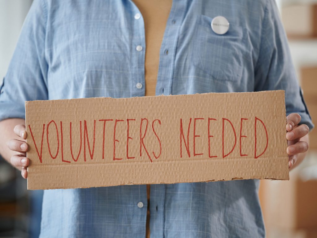 Woman in a blue shirt holds a handwritten cardboard sign that reads "Volunteers Needed"